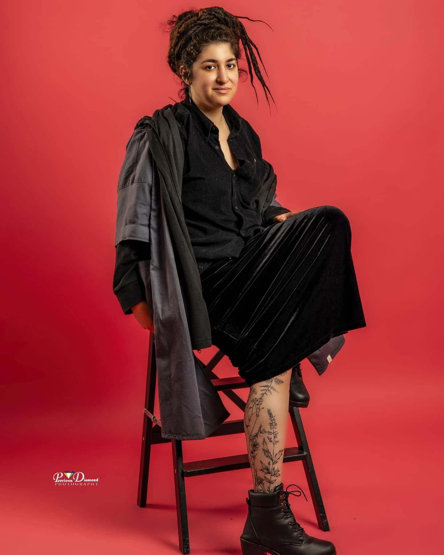 Full-length studio portrait on a wooden chair against a red backdrop, dressed in black with lace-up boots