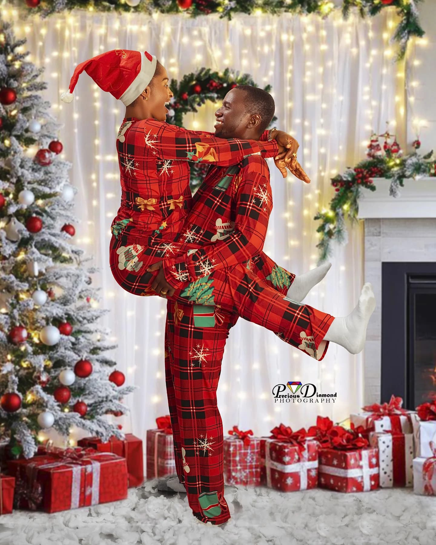 A couple in matching red tartan on a Christmas set, one leaping into the other’s arms beside a snowy tree and wrapped presents