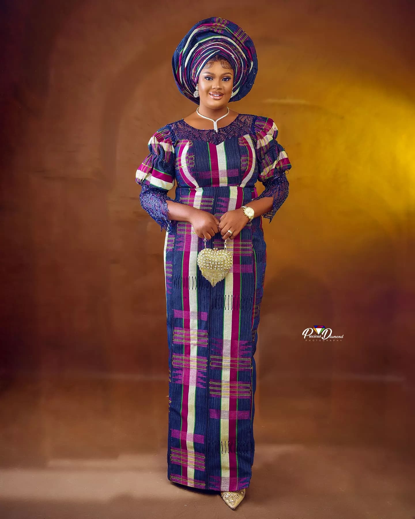Full-length portrait in traditional attire: a striped purple and blue outfit with a matching gele, against a warm golden backdrop
