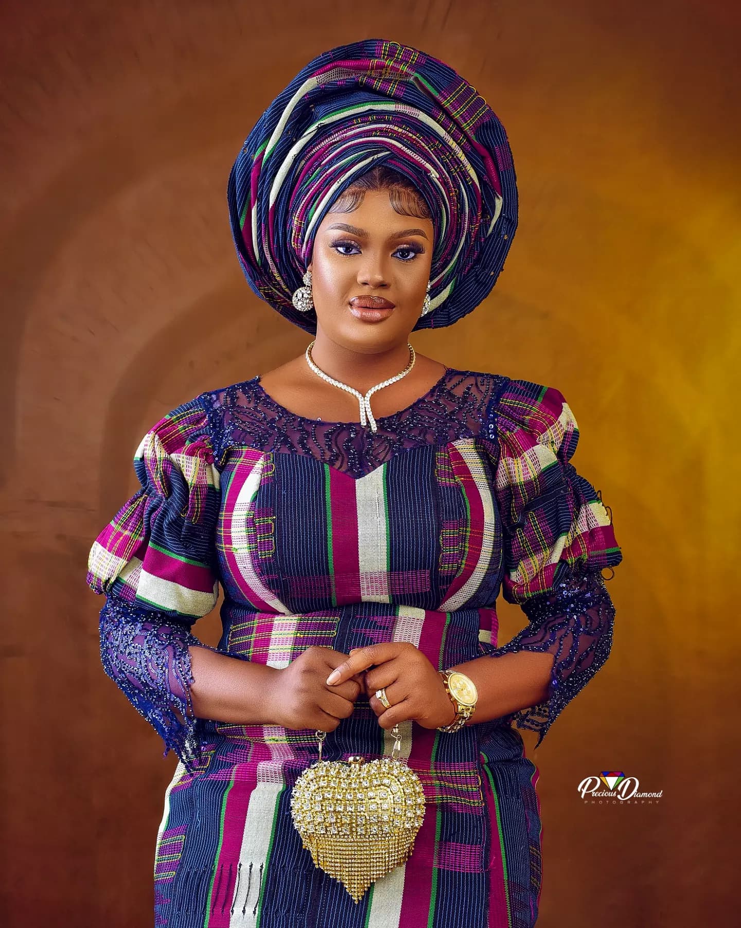 Smiling portrait in a purple and blue gele and matching outfit, holding a gold heart-shaped clutch