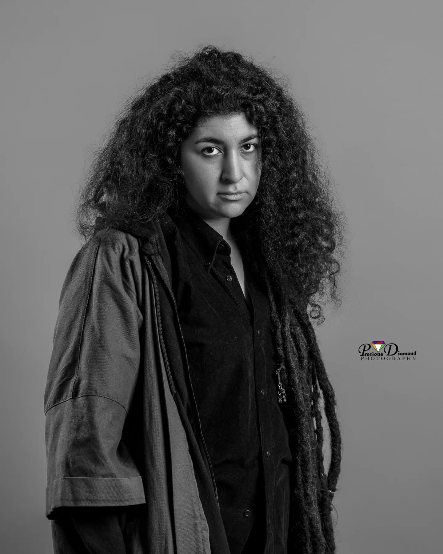 Black and white studio portrait, hands in the pockets of a long coat, looking straight at the camera