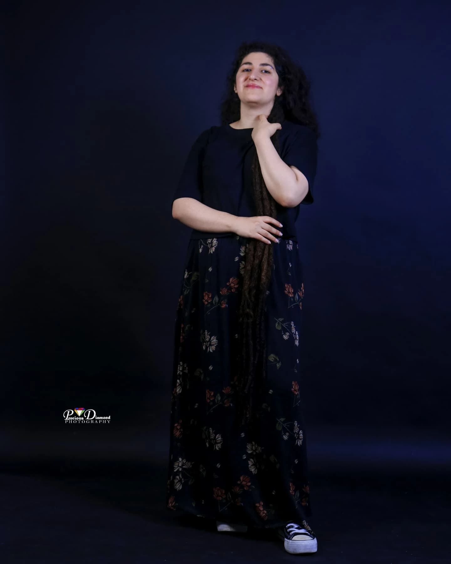 Full-length portrait on a navy backdrop in a black top and floral skirt, smiling, one hand at the chest