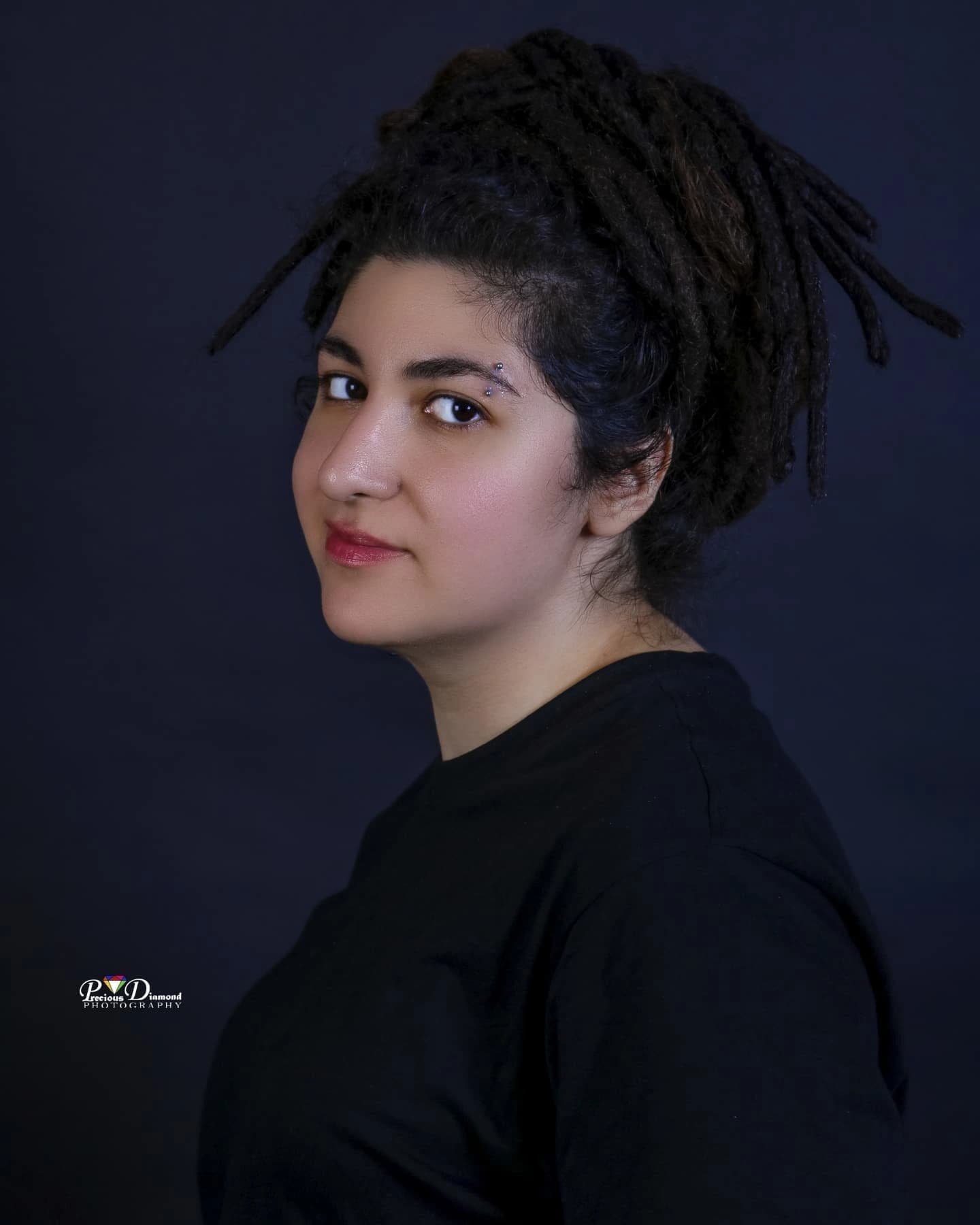Portrait on a navy backdrop looking back over one shoulder, locs gathered high on the head