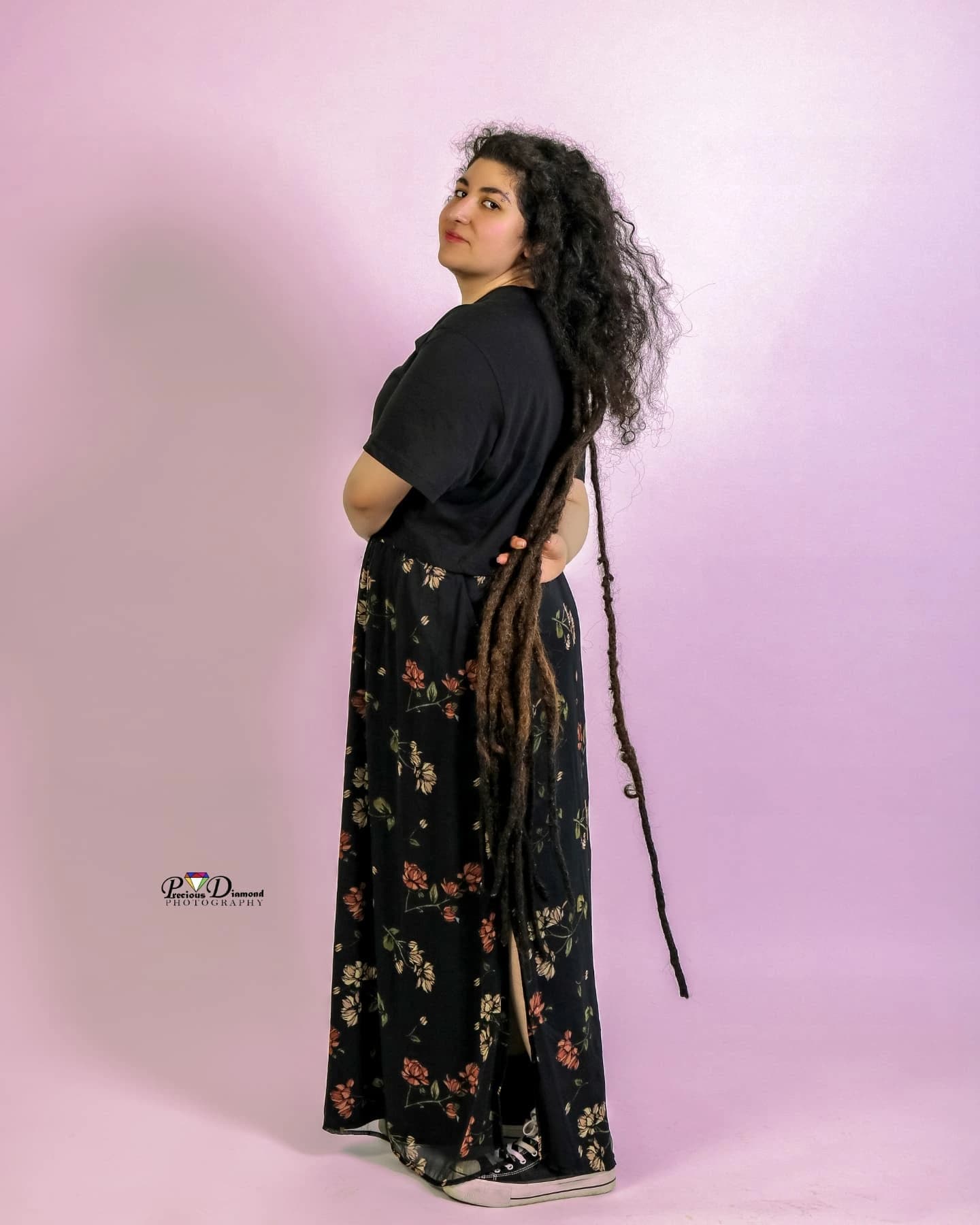Full-length portrait on a soft pink backdrop, looking back over one shoulder, with floor-length locs and a floral skirt