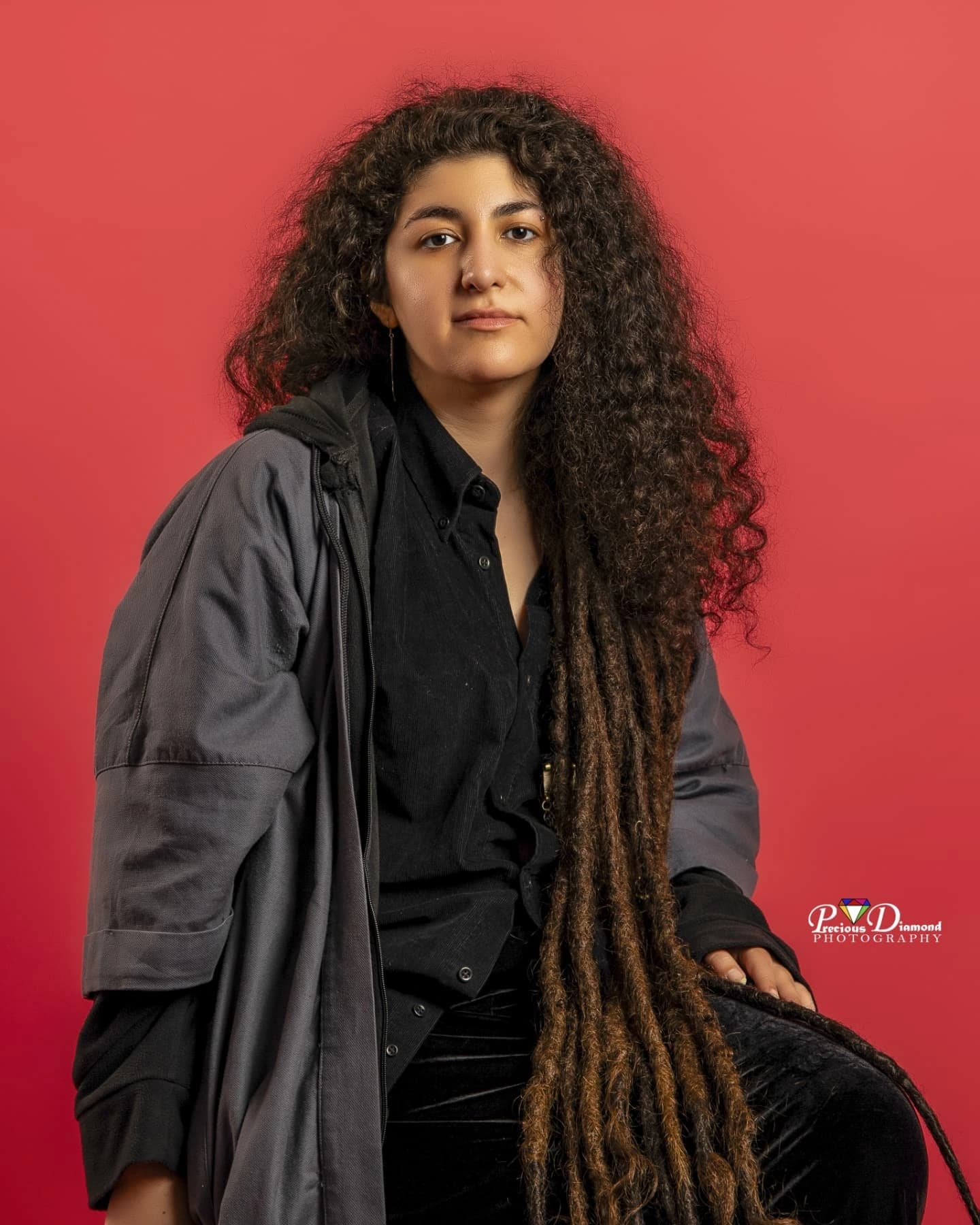 Seated studio portrait on a red backdrop, in an oversized grey coat with very long locs falling to the lap
