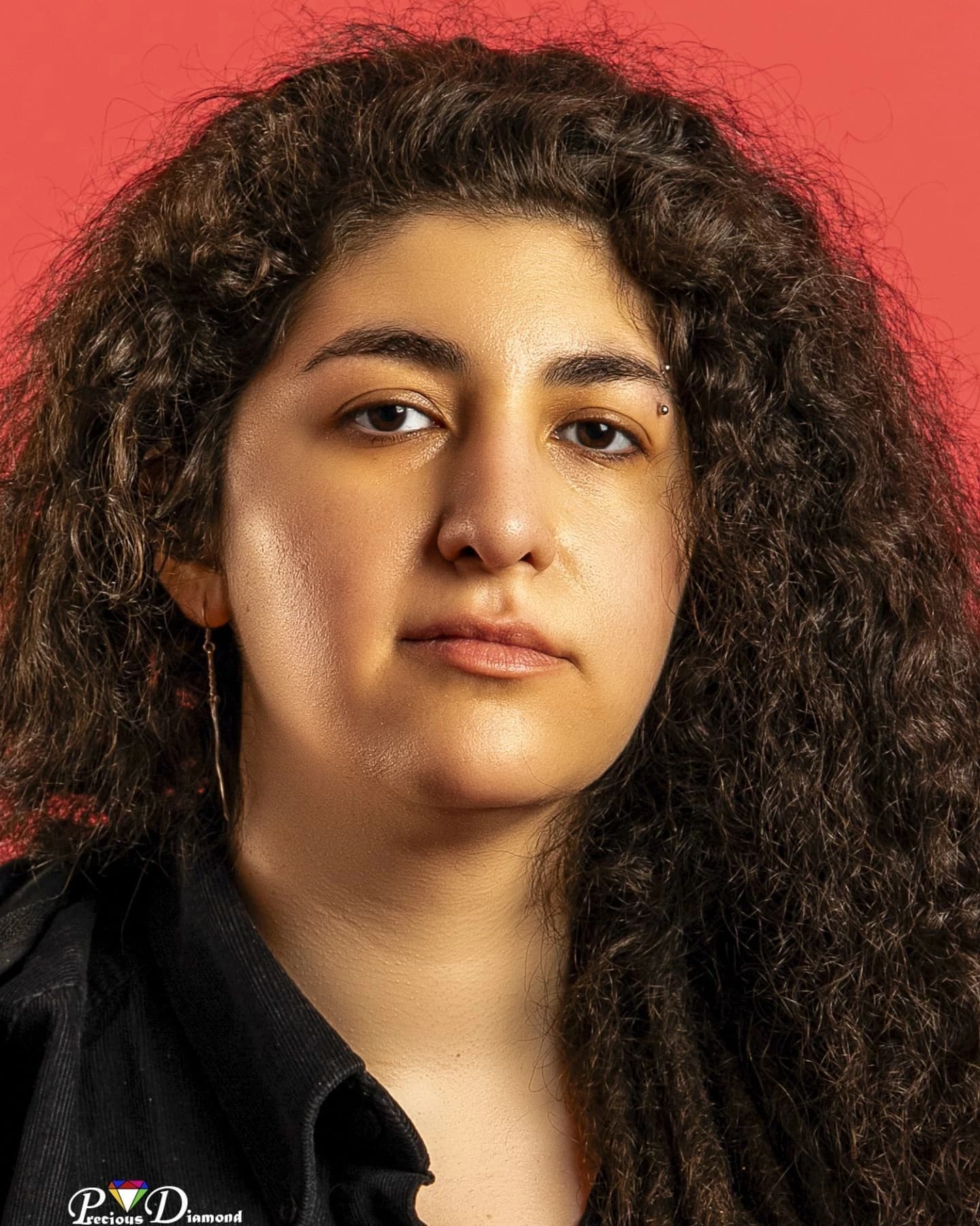 Close studio portrait against a red backdrop: long dark curls, a black collar and a calm, direct gaze