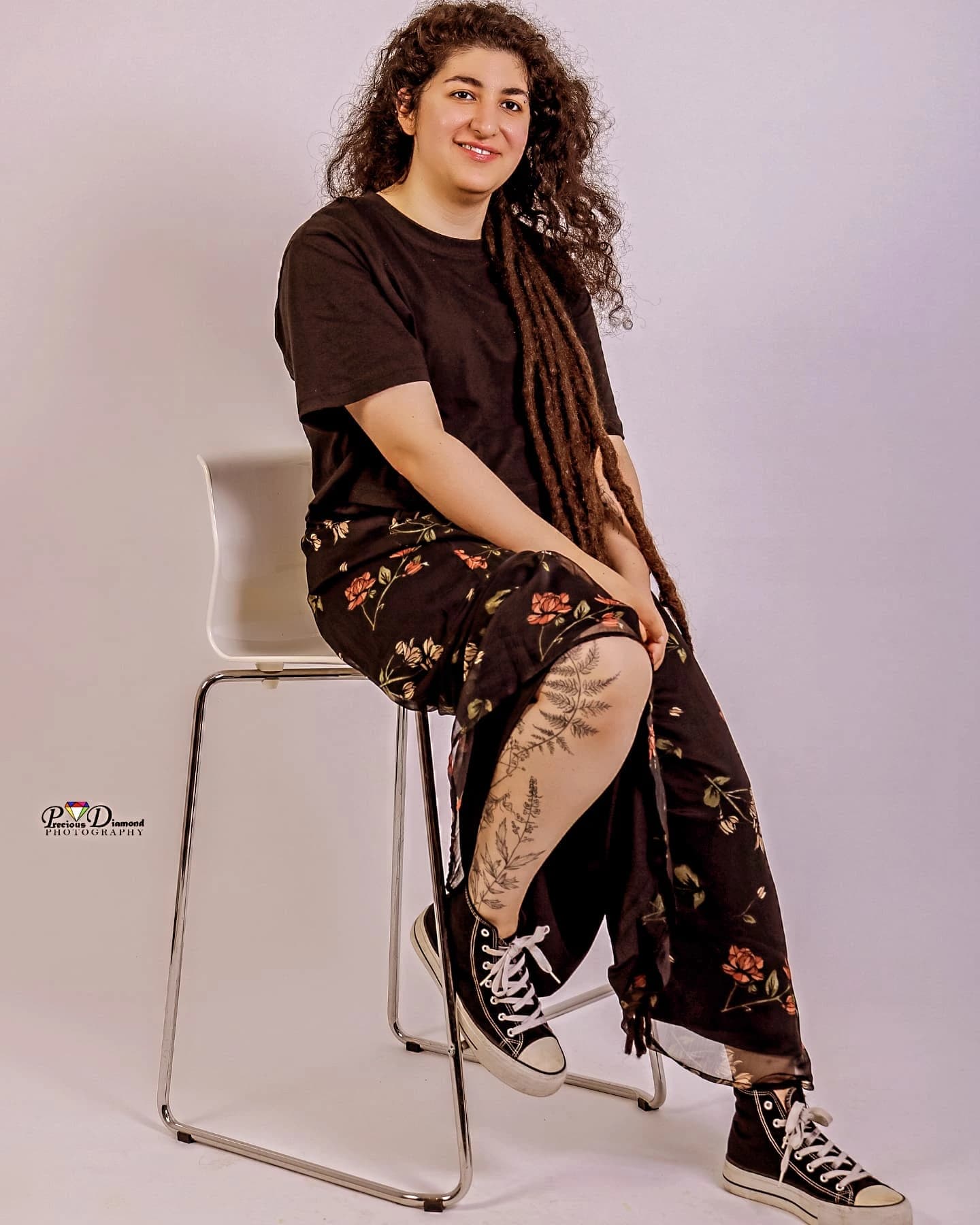 Smiling portrait seated on a tall stool against a pale backdrop, in a floral skirt and trainers